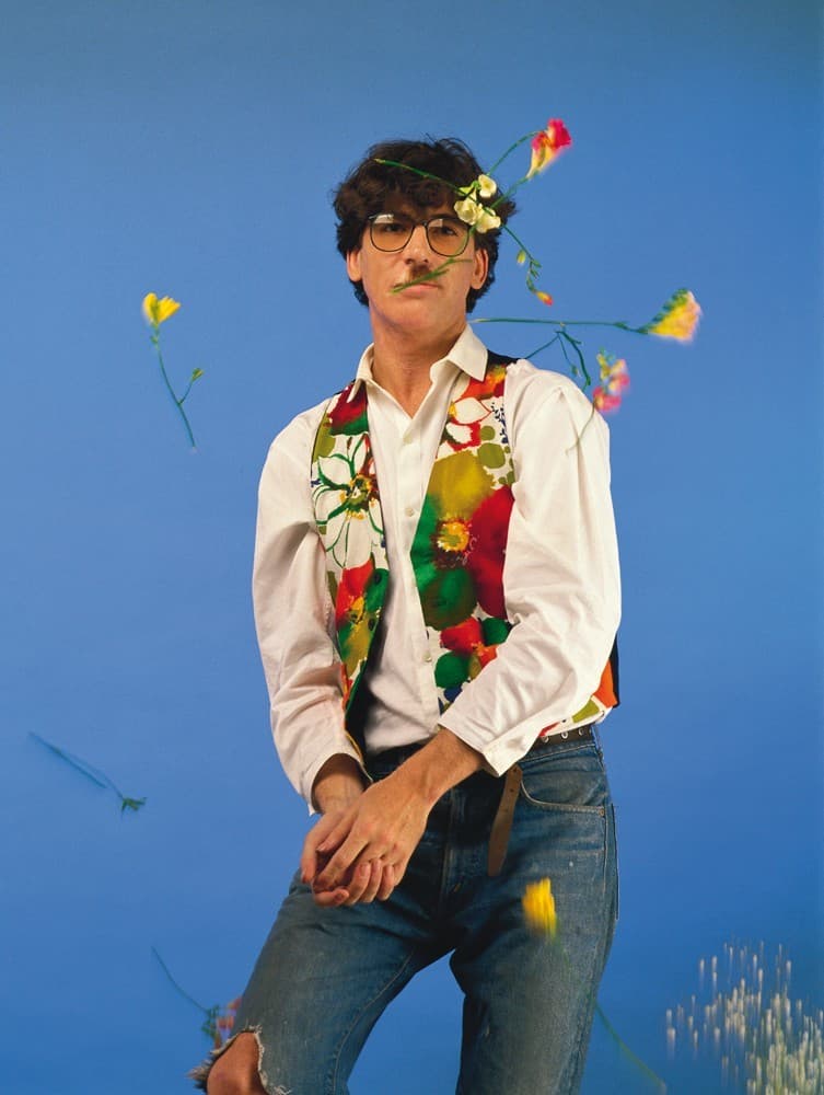 Persona con gafas, camisa blanca y flores de colores alrededor del torso frente a un fondo azul.